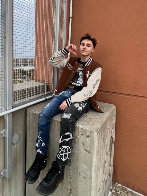 Young man with black hair sitting on a concrete block overlooking a city. He's wearing jeans, black boots, and a brown letterman jacket